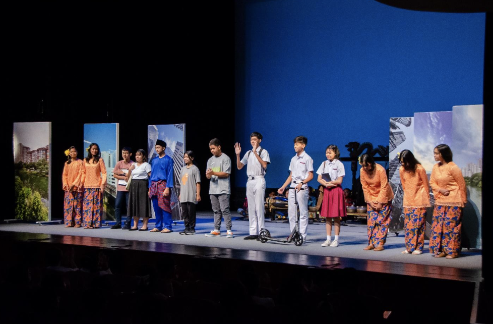 A group of children and adults perform on a stage, some in orange traditional outfits, against a blue backdrop.