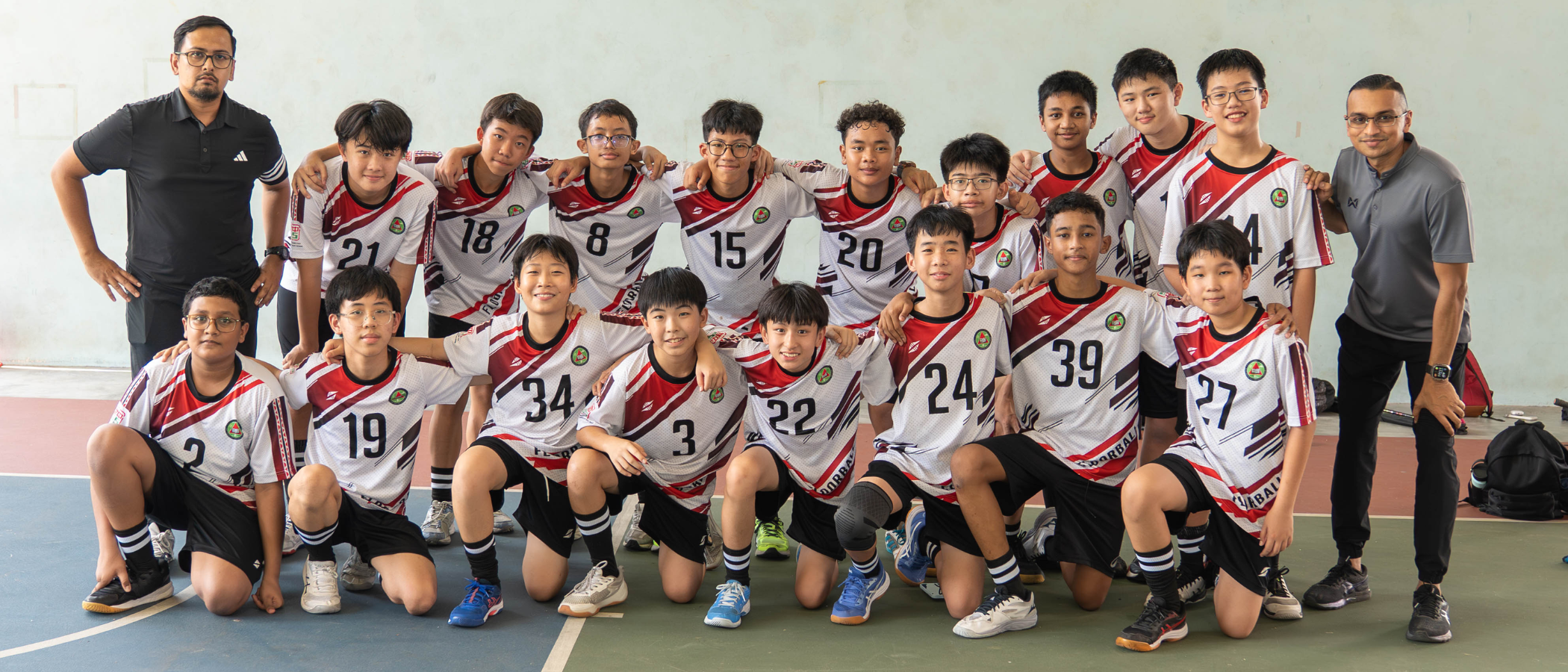 A youth sports team in white and red numbered jerseys poses with two adult coaches on an indoor court.