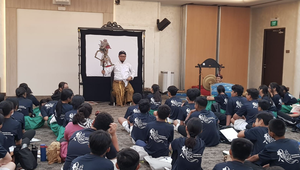 A performer holds a traditional wayang shadow puppet before a large group of students seated on the floor.