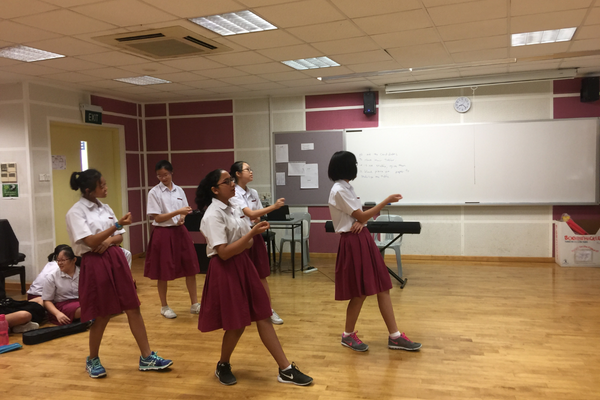 Several schoolgirls in white shirts and maroon skirts practice a dance routine in a classroom with a whiteboard.
