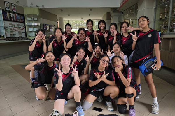 A group of teenage girls in black sports uniforms posing together, many making peace signs with their hands.
