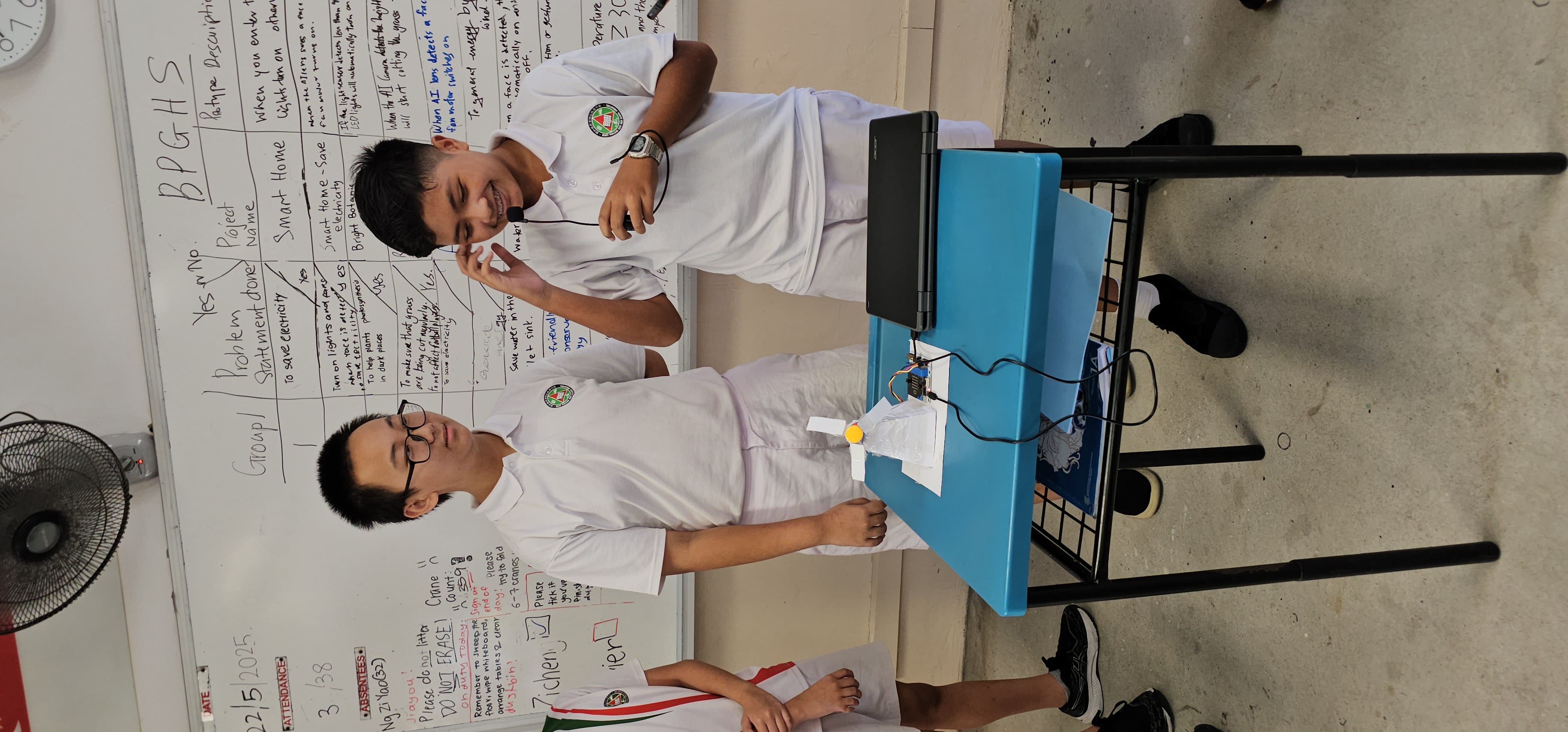 Two male students in white uniforms present a Smart Home electronics project on a blue table before a whiteboard.