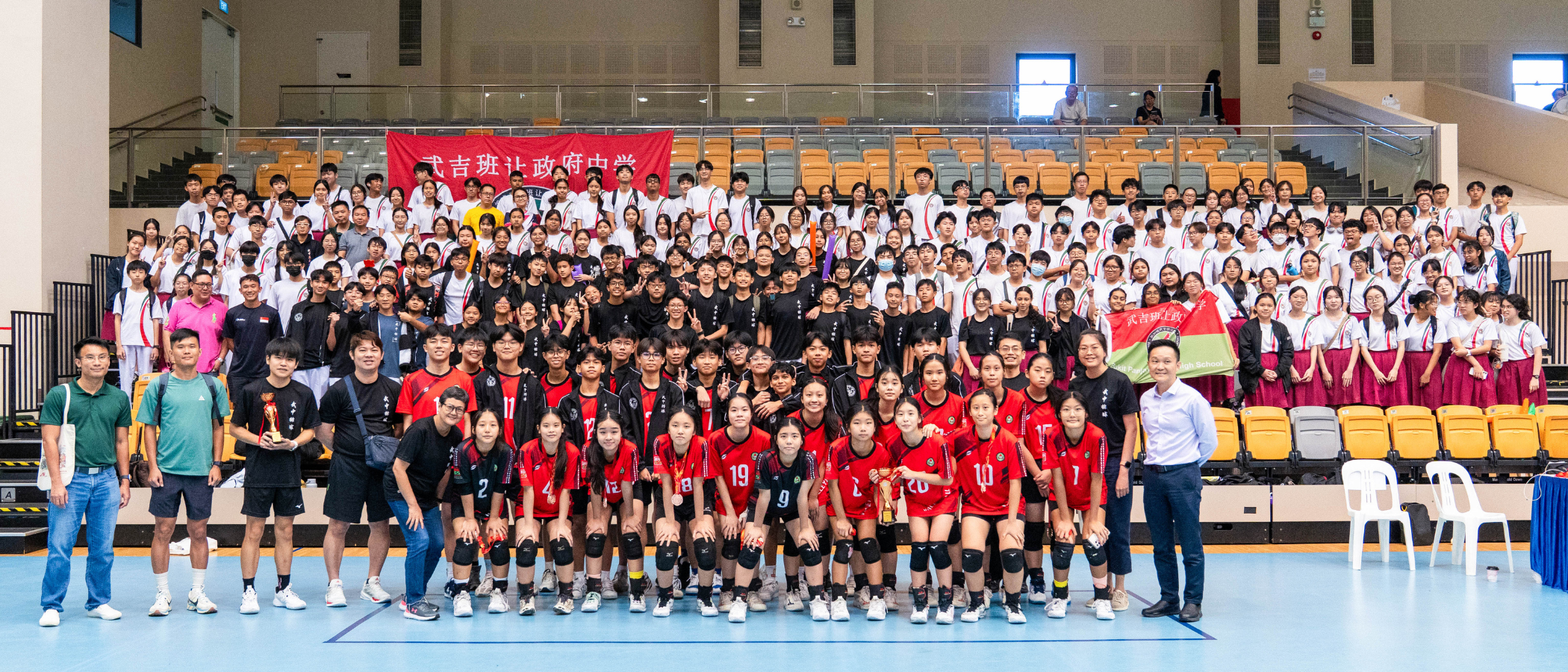 Large group of students posing in an indoor sports hall, front row in red volleyball jerseys holding trophies.