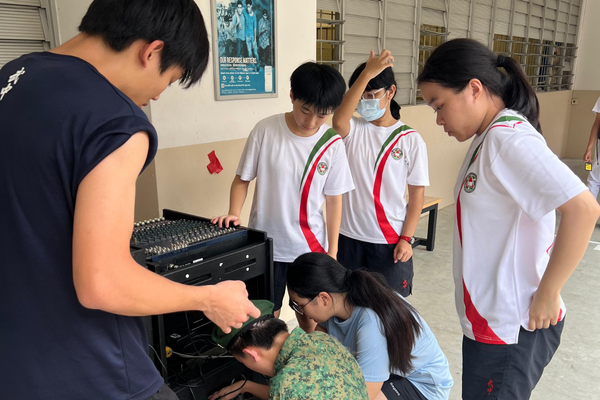Several students in white uniforms gather around adults working on audio mixing equipment indoors.