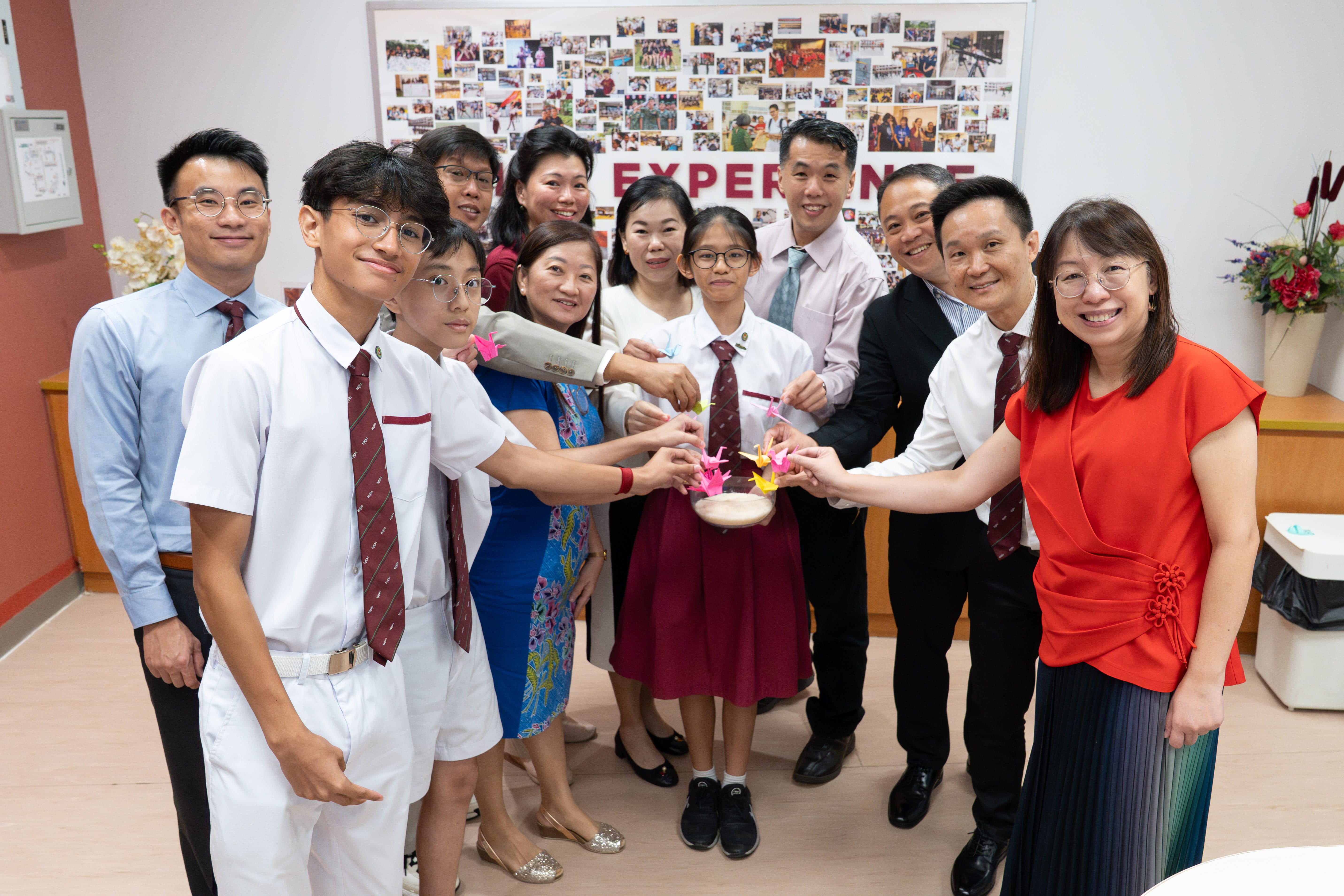 A group of students in uniforms and adults smile while placing colorful origami cranes into a bowl indoors.