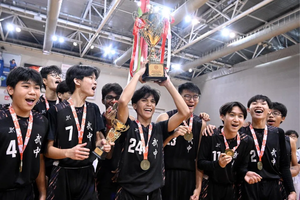 Young basketball players in black jerseys with medals celebrate as one raises a championship trophy overhead.
