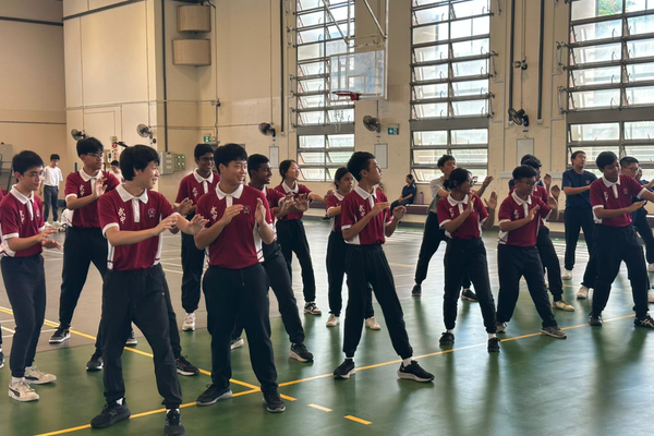 Students in matching red polo shirts practice martial arts or dance moves together in a large indoor gymnasium.