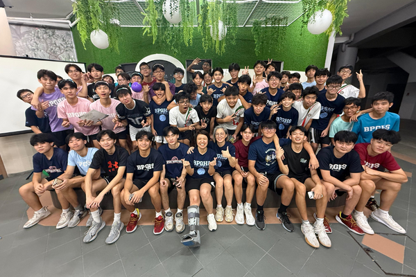 A large group of young students wearing matching school shirts pose together indoors in front of a green wall.