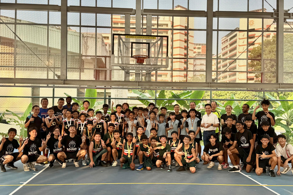 A large group of youth basketball players and coaches pose together on an outdoor court under a hoop.