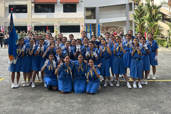 A large group of girls in blue uniforms and white gloves pose outdoors making peace signs near a school building.
