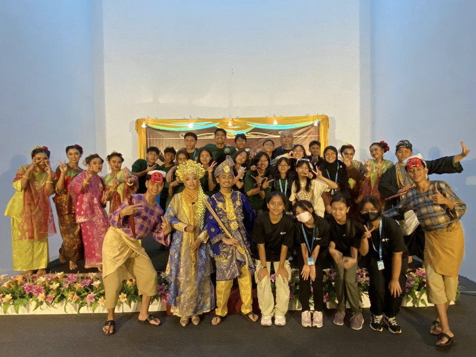 A large group of people posing together, some in colorful traditional Indonesian costumes, others in casual clothing, on a decorated stage.