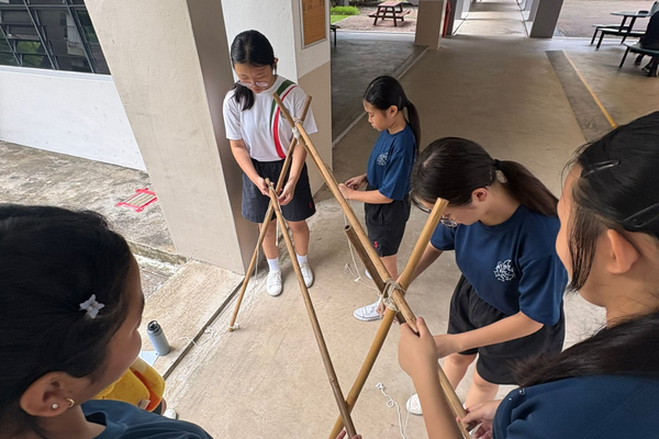 Four students in school uniforms work together outdoors to build a tripod structure using bamboo poles and rope.