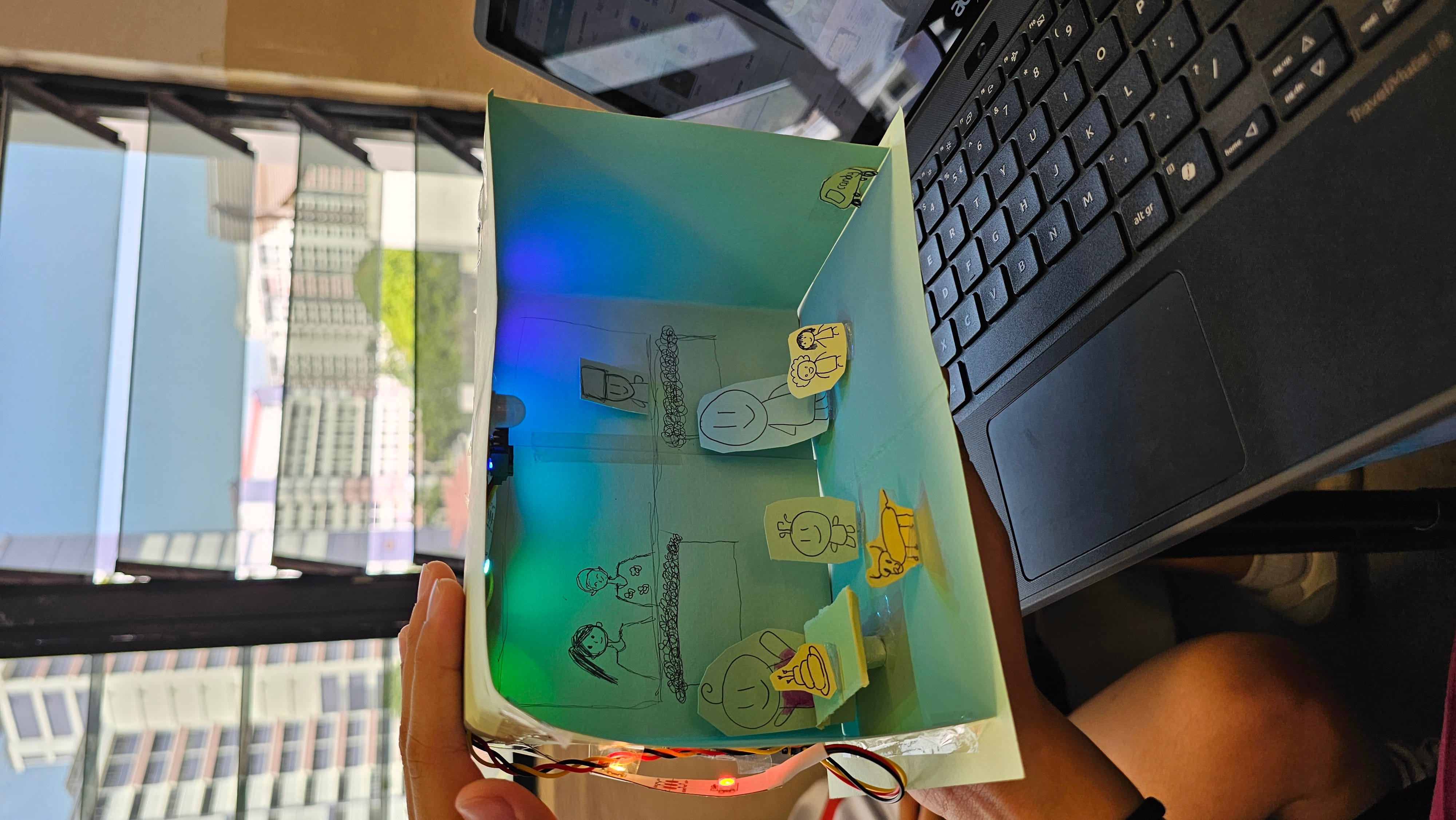 A hand holds a paper diorama with hand-drawn stick figures and LED lighting, resembling a miniature indoor scene.
