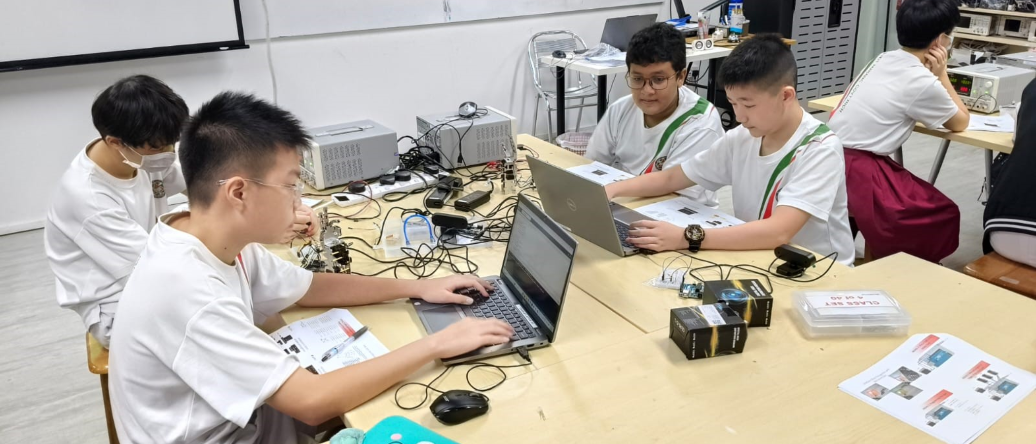 Four students in white shirts work on laptops and electronics at tables in a classroom lab with cables and equipment.