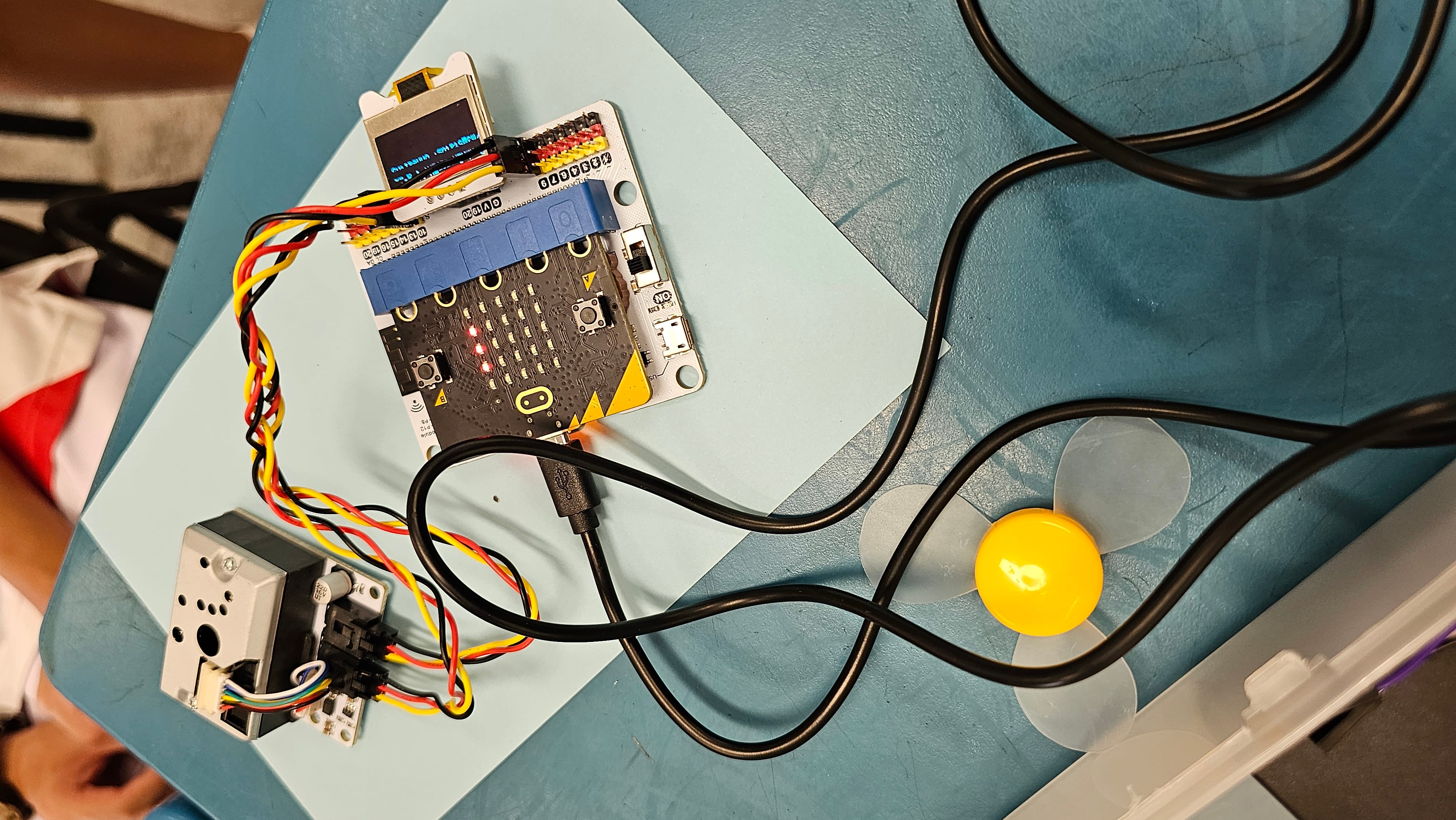 A micro:bit circuit board with colorful wires, a sensor module, small fan, and USB cable on a blue table.