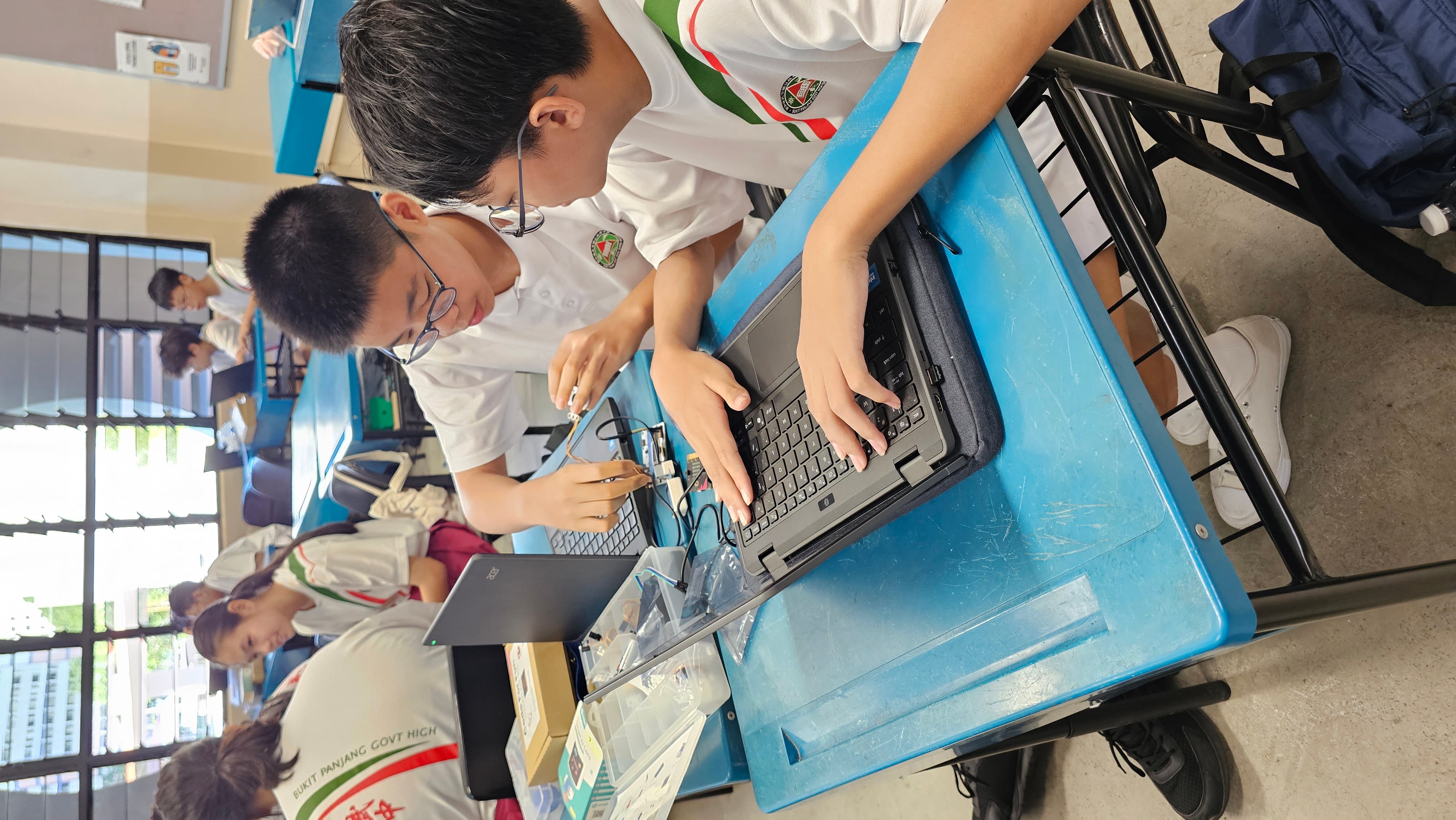 Two school students in white uniforms work together on a laptop and electronic components at a blue desk in a classroom.