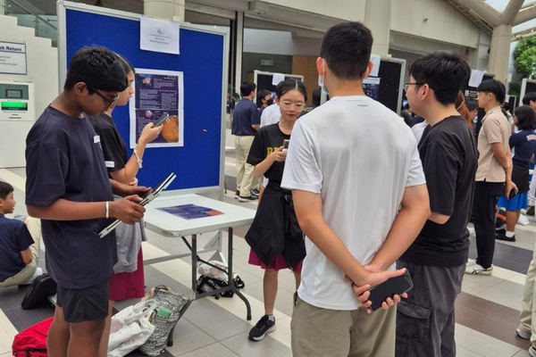 Students gather around a display board at an indoor fair, holding phones and papers while discussing a project.