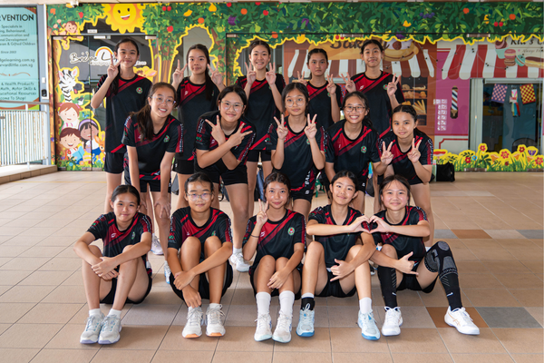 A group of young girls in black and red sports uniforms pose together in three rows, making peace signs.