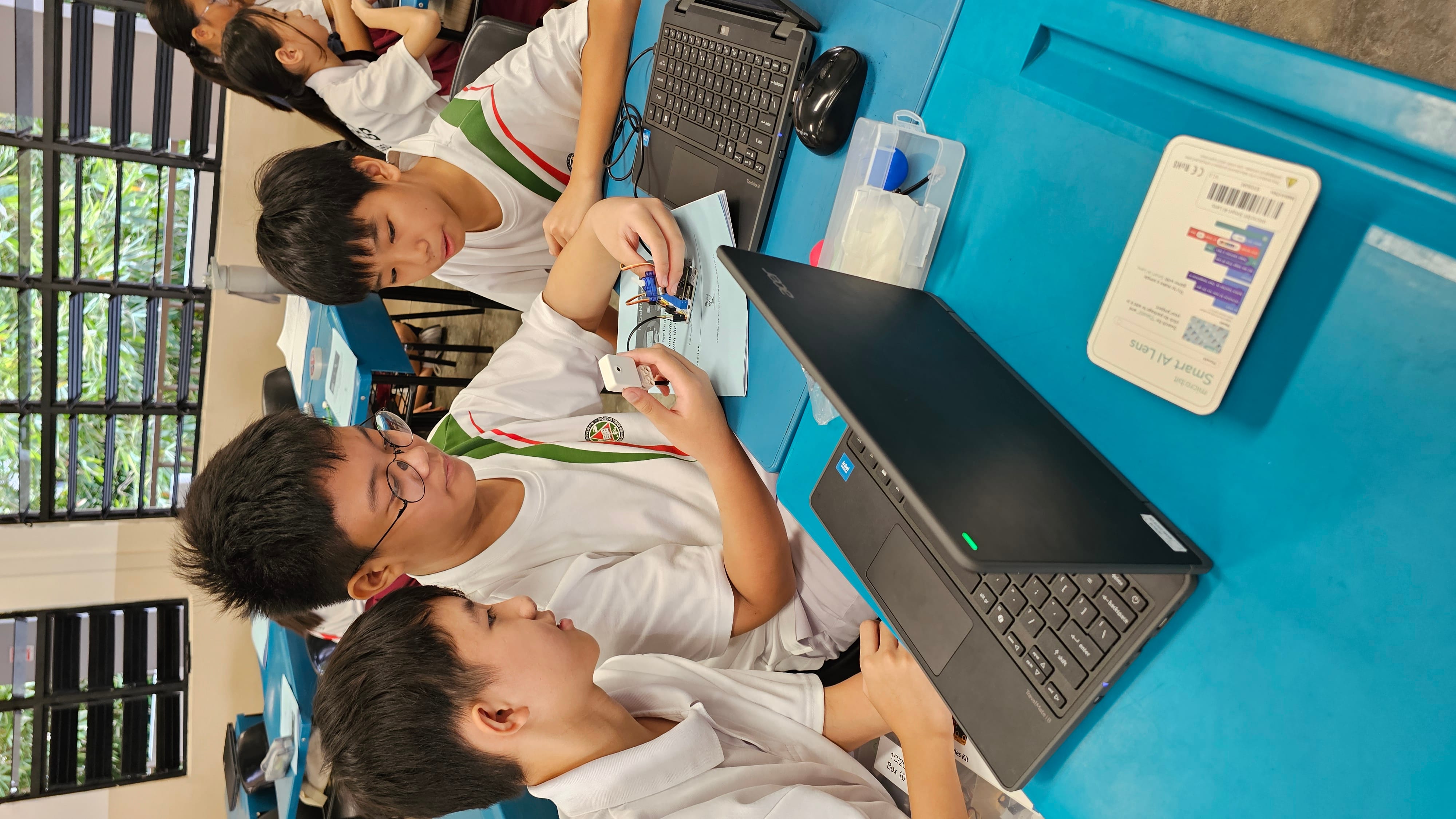 Three school boys in white uniforms work together on an electronics project at a blue desk with laptops nearby.