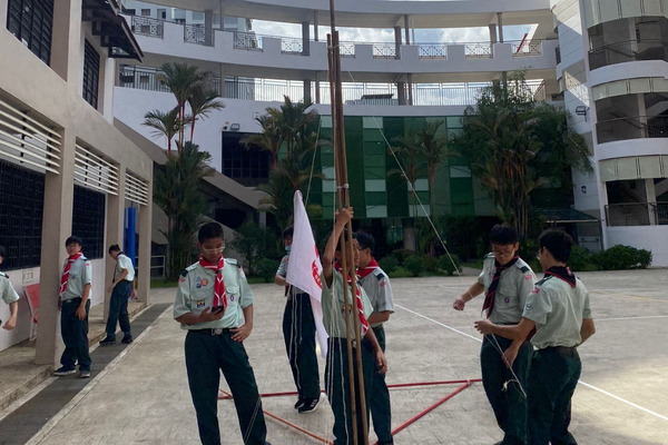 Boy scouts in uniform raising a white flag on a tall pole in a school courtyard.