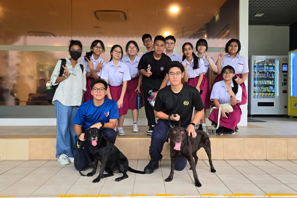 A group of students in school uniforms pose outdoors with two black dogs, with a vending machine visible on the right.