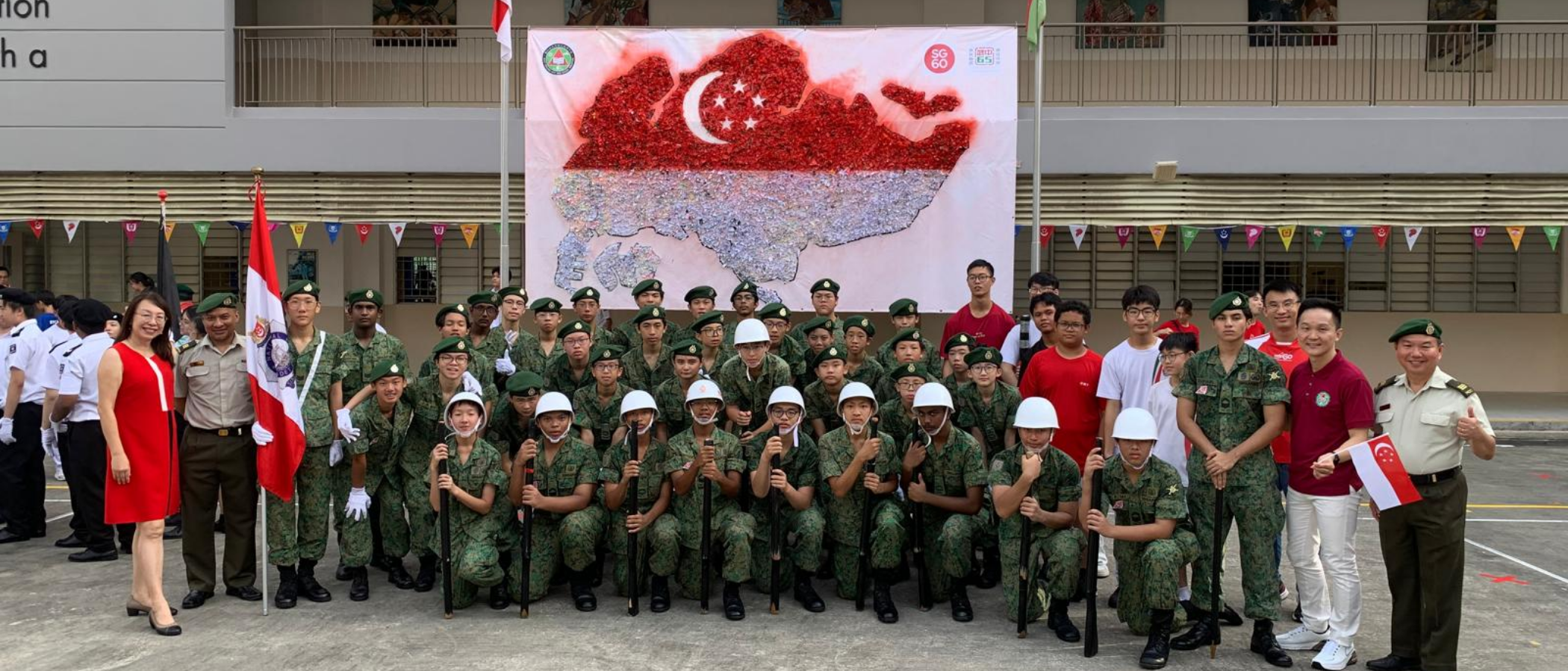 Group of youth cadets in green camouflage uniforms posing before a Singapore flag map display at an SG60 event.