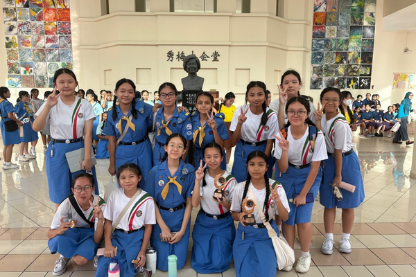 A group of girls in blue scout uniforms pose with peace signs in front of a bust statue indoors.