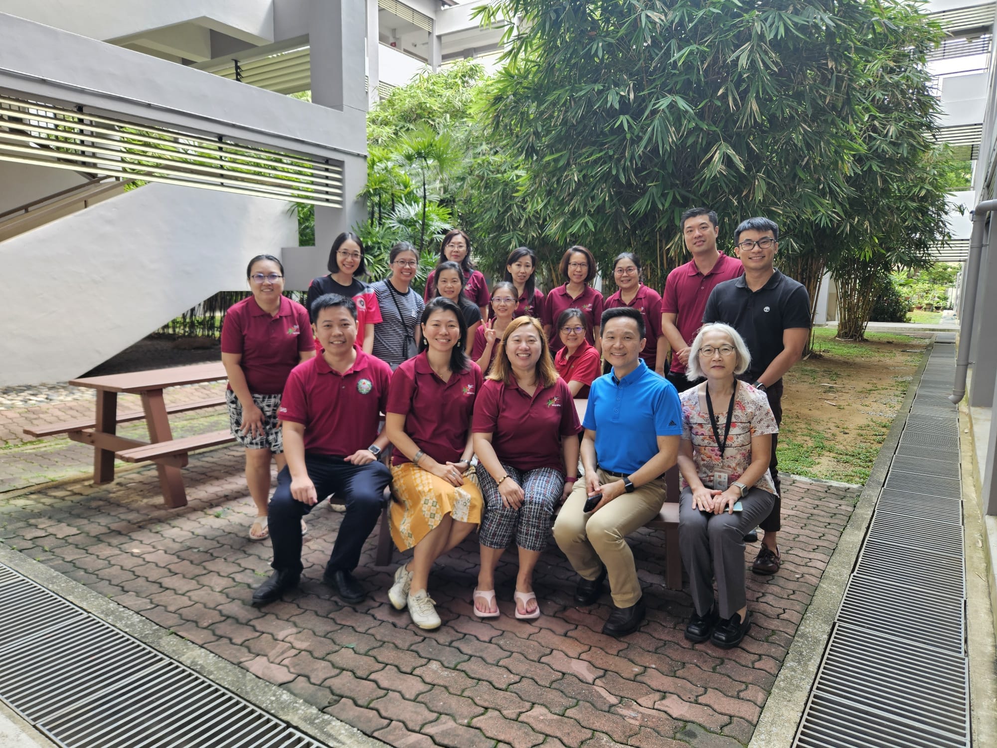 A group of about 18 people posing outdoors on a brick courtyard, most wearing matching dark red polo shirts.