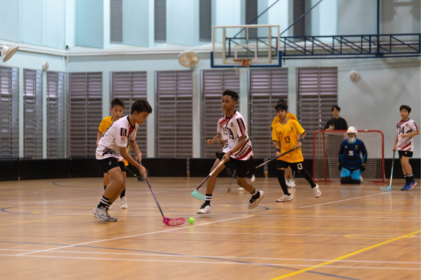Boys playing floorball indoors, competing with sticks near a small ball, with a goalkeeper and goal in the background.