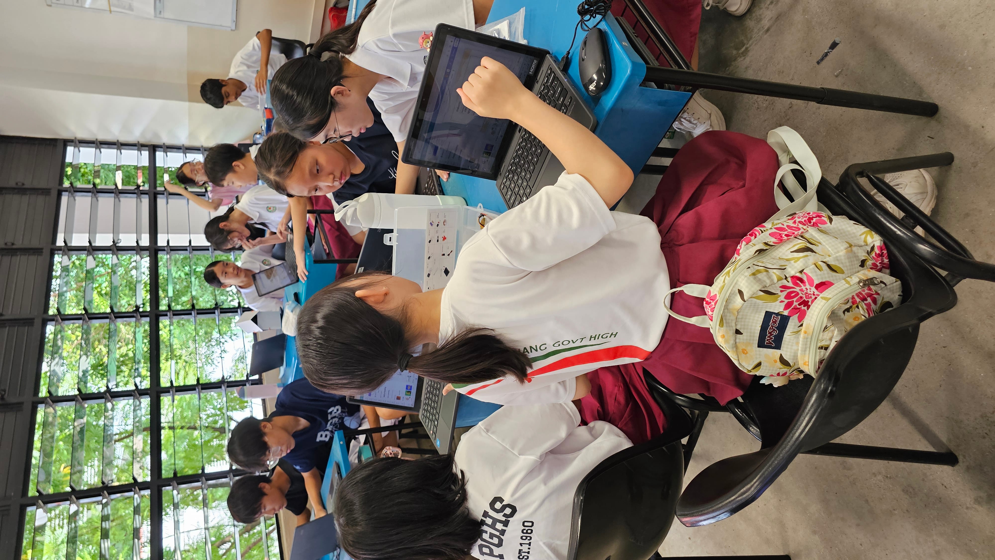 Students in school uniforms sit at blue tables working on laptops in a classroom with large louvered windows.