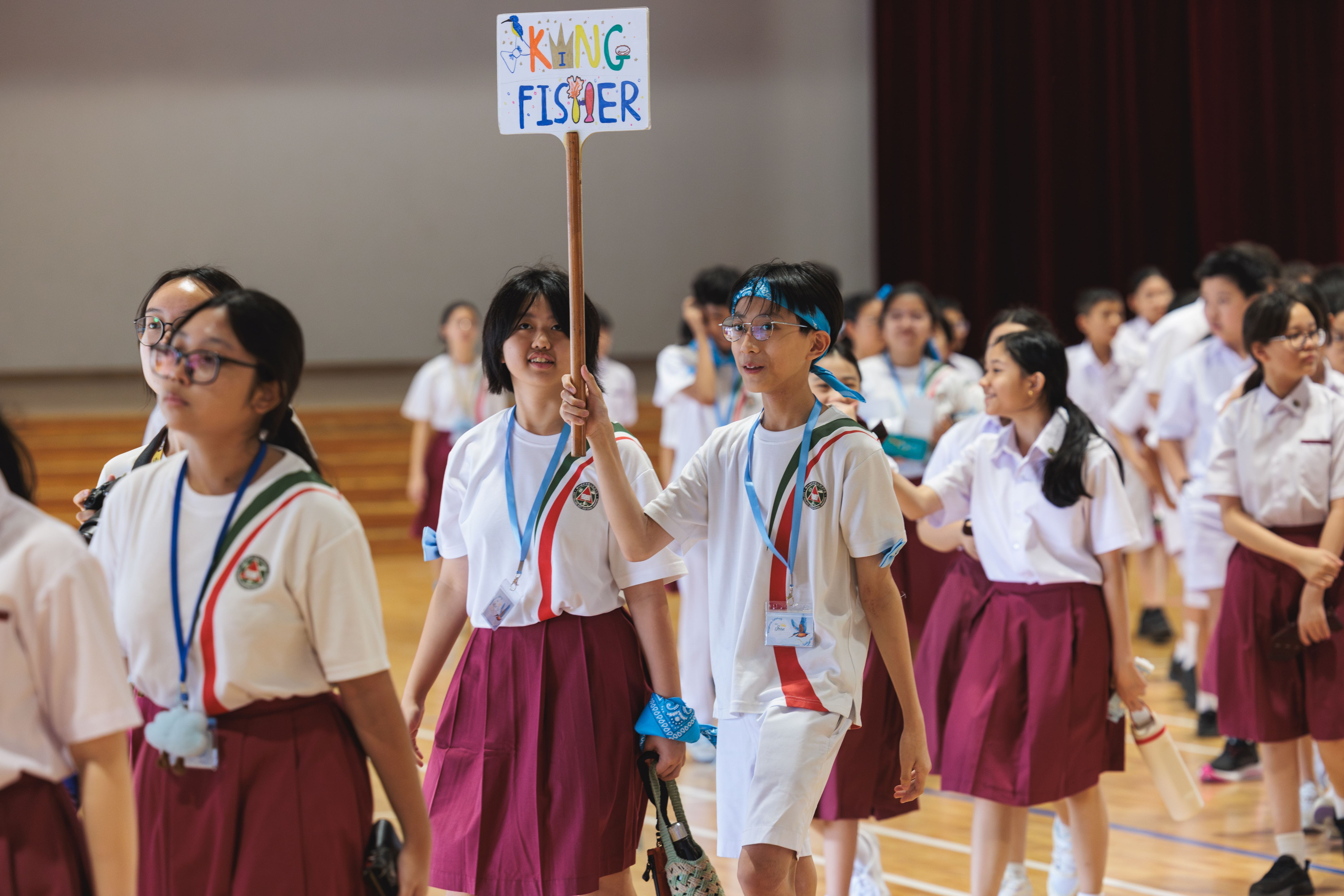 Students in white and maroon uniforms walk in a hall as one holds a colorful sign reading "Kingfisher."