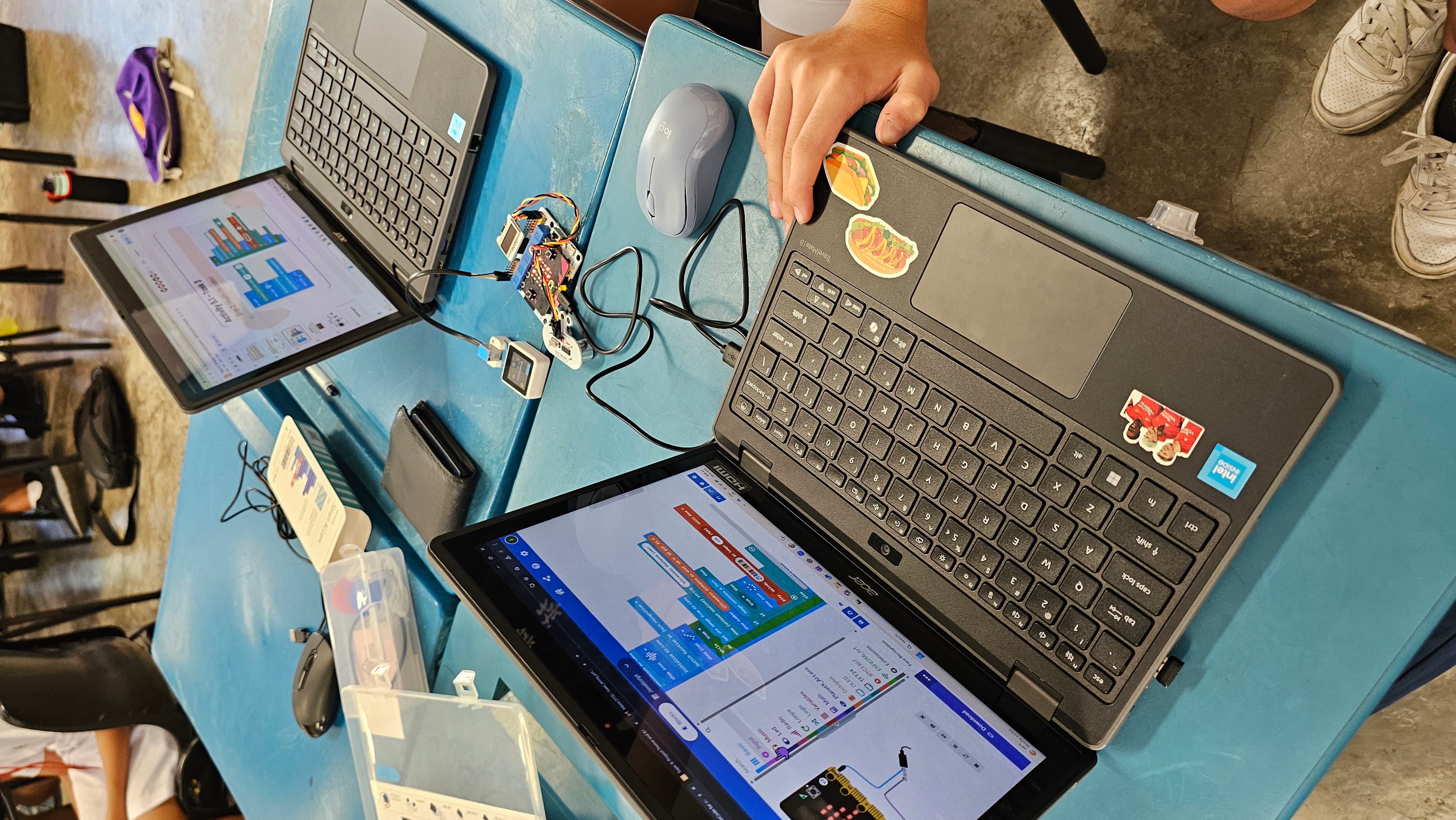 Two laptops on a blue desk showing block-based coding, with a circuit board and mouse nearby, and a hand on one laptop.
