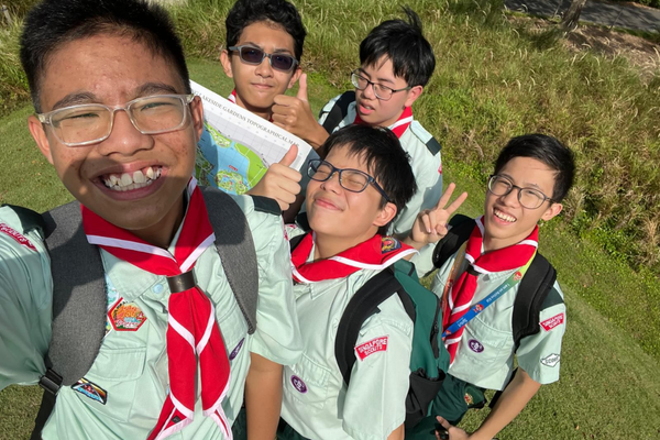 Five young scouts in green uniforms with red neckerchiefs smile and pose for a selfie outdoors.