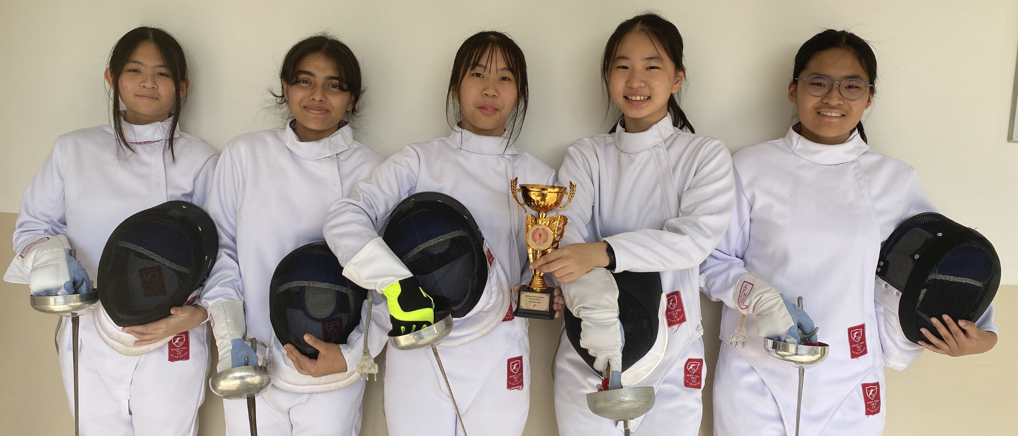Five girls in white fencing jackets hold helmets and swords, with one holding a gold trophy cup.
