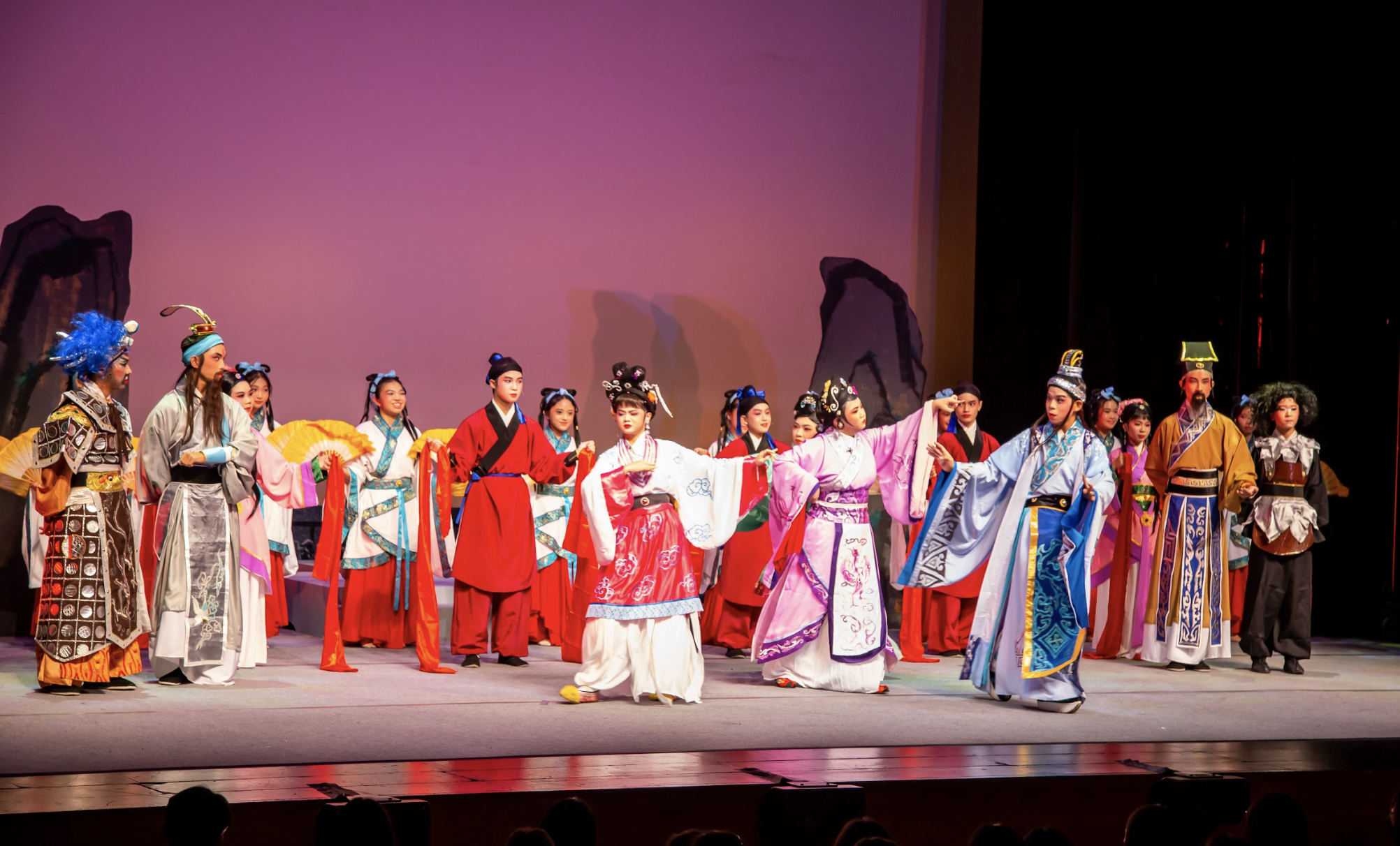 A large cast in colorful traditional Chinese opera costumes performs on a stage with a pink backdrop.