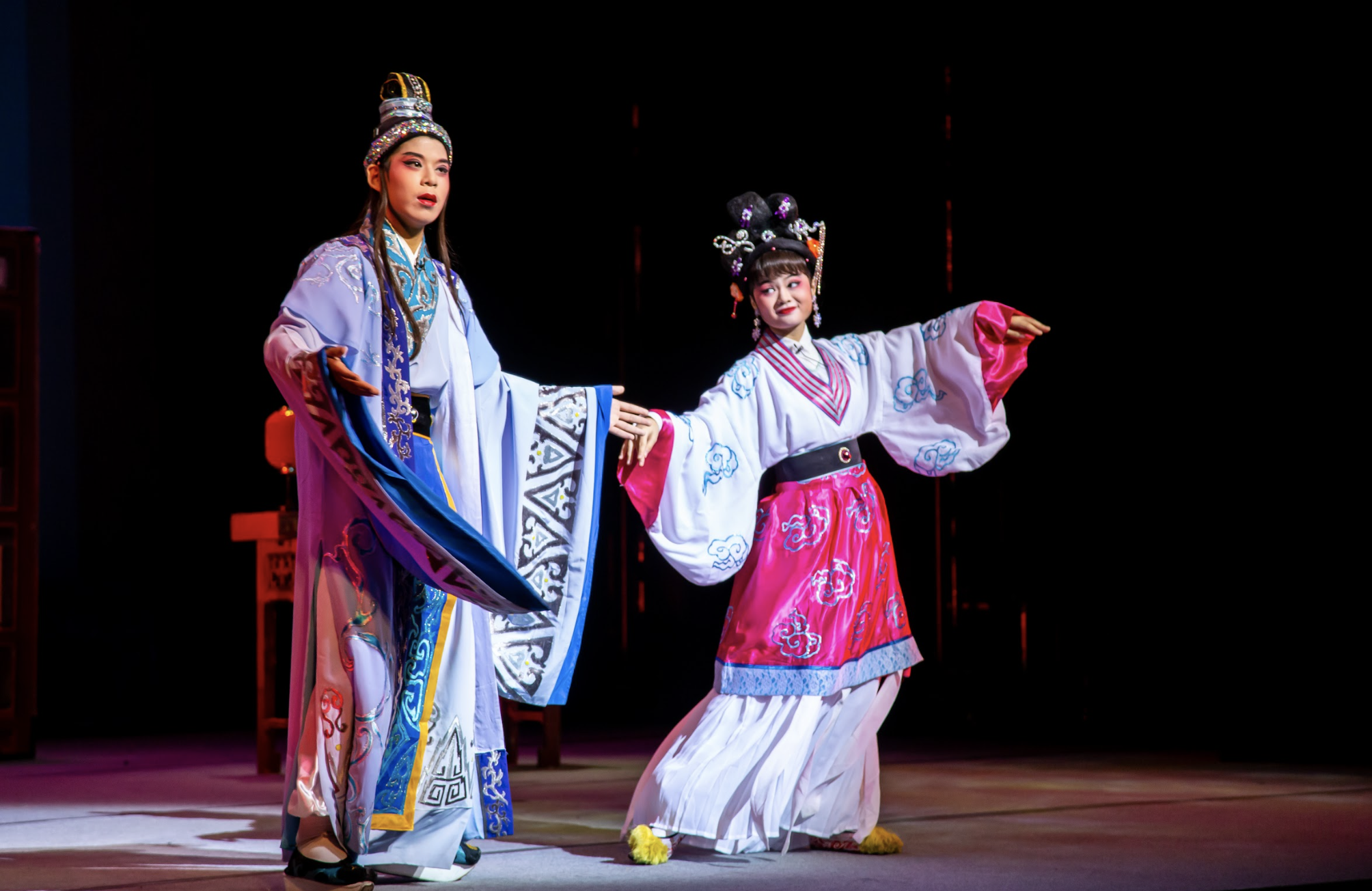 Two performers in traditional Chinese opera costumes and headdresses pose dramatically on a dark stage.