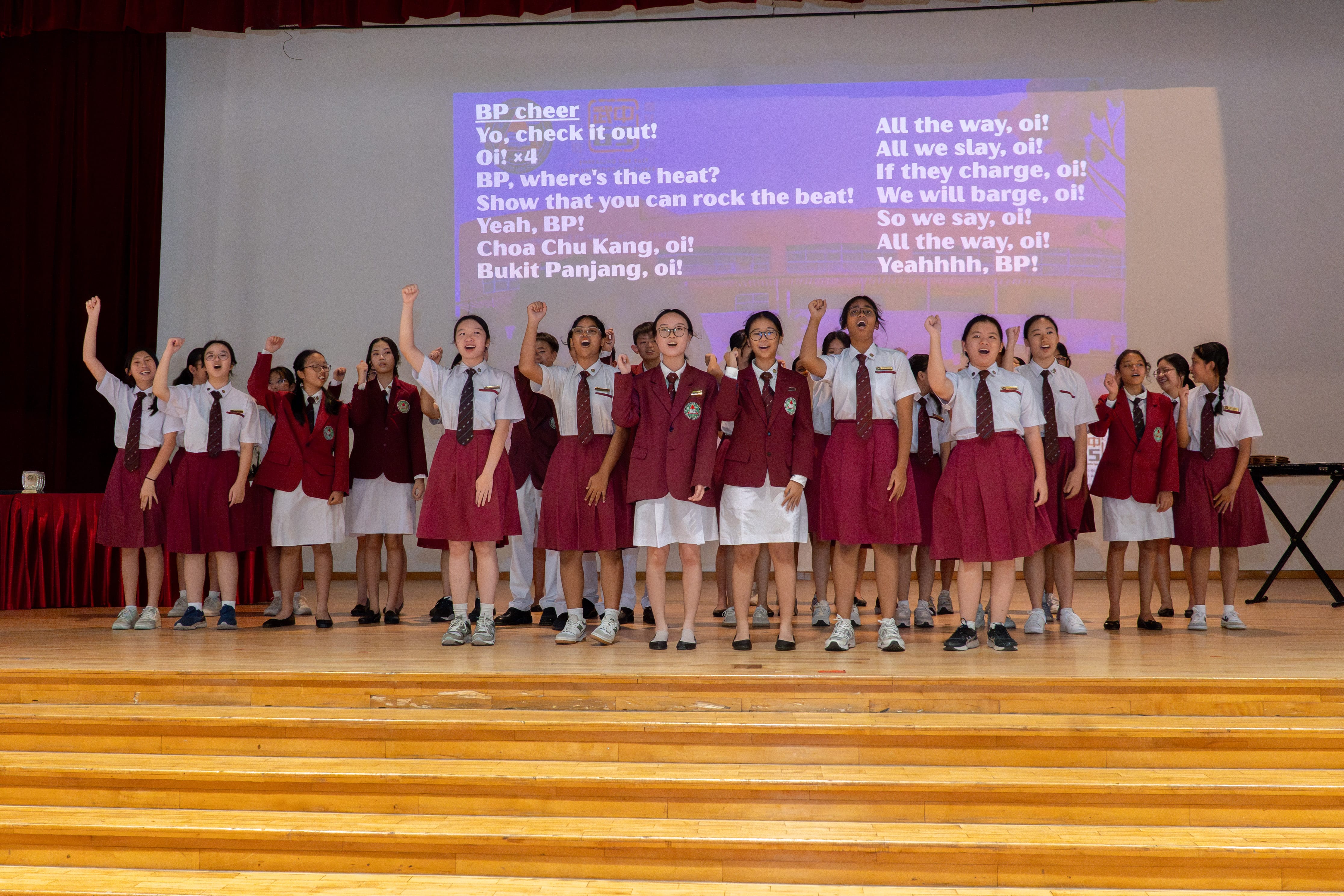 Students in red and white uniforms perform a school cheer on stage with cheer lyrics projected behind them.