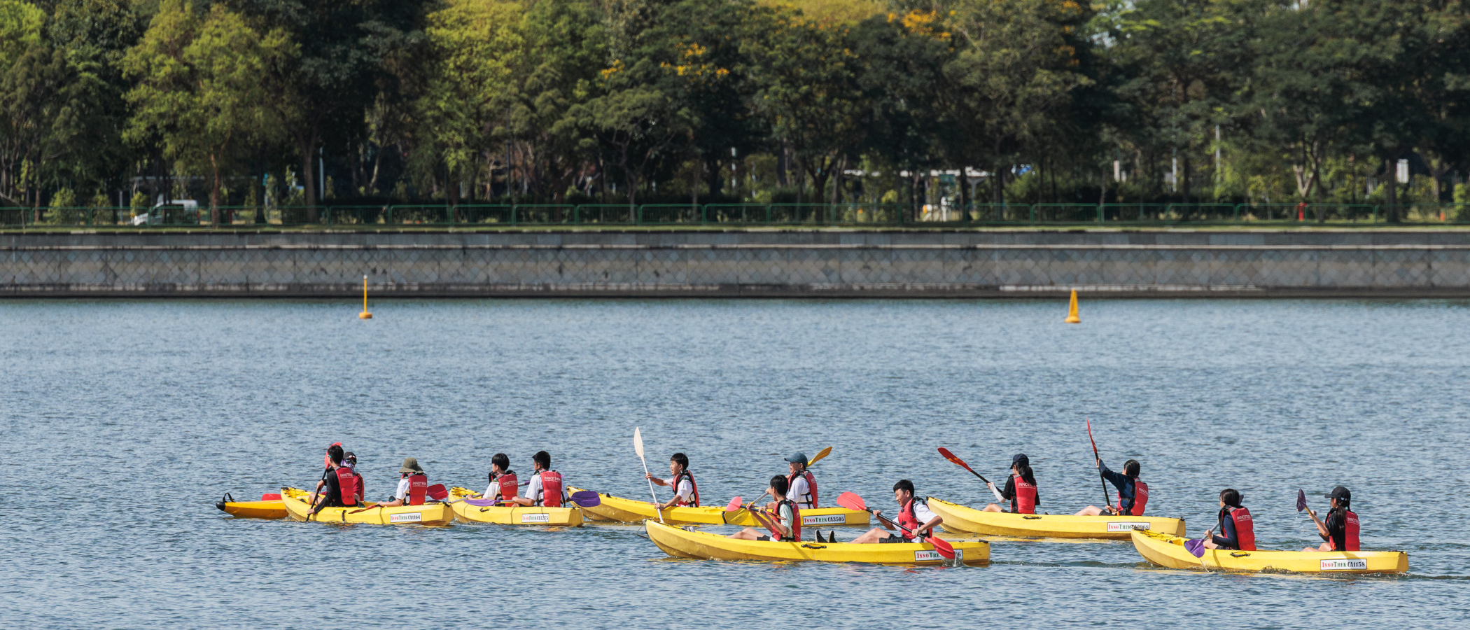 A group of people in red life vests paddling yellow kayaks on a calm lake with trees in the background.