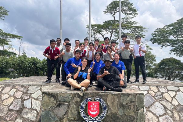A group of students posing outdoors near a stone monument with a Singapore crest, flagpoles, and trees in the background.