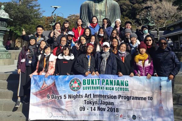A group of students pose before a large Buddha statue holding a Bukit Panjang Government High School Tokyo 2018 trip banner.
