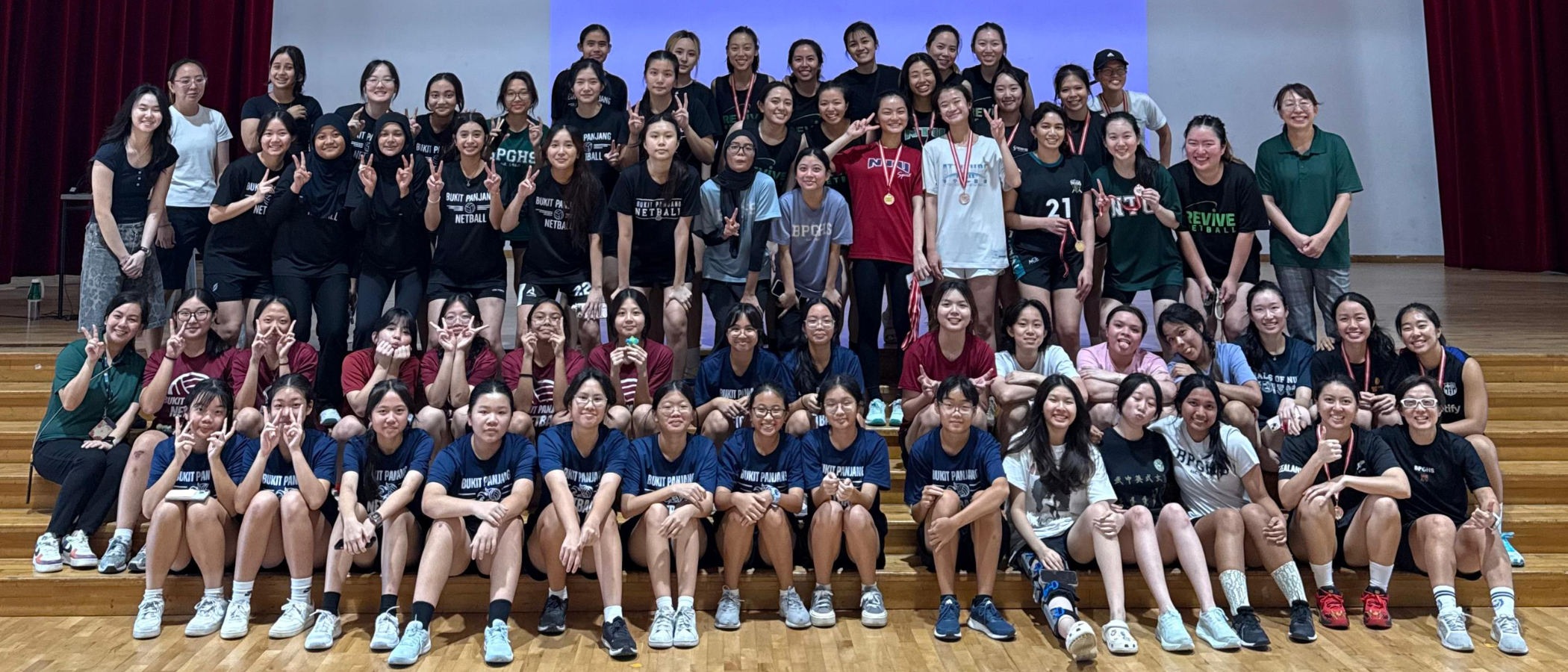 Large group of young female netball players posing together on a wooden gym floor, some wearing medals.