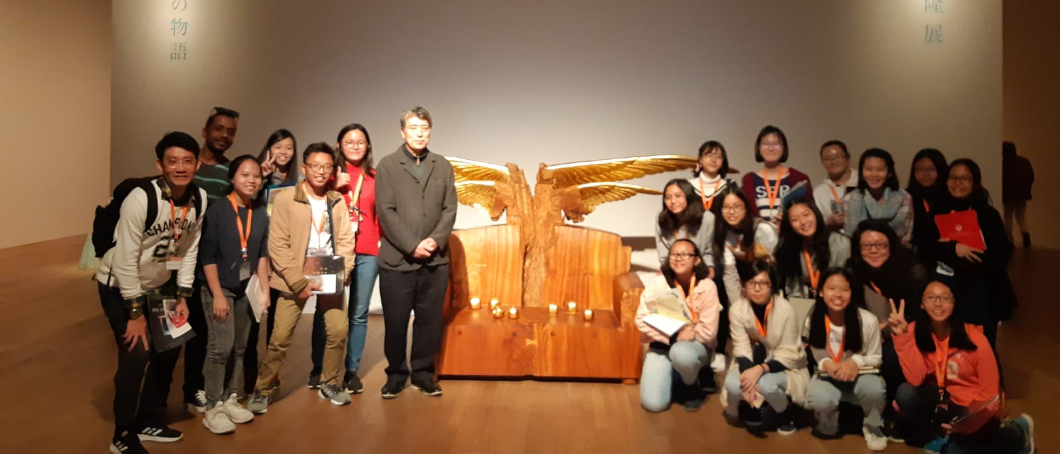 A group of about 20 young people with orange lanyards pose beside a golden winged wooden sculpture in a museum.