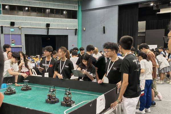 Students in black shirts gather around a table with small robots at an indoor robotics competition.