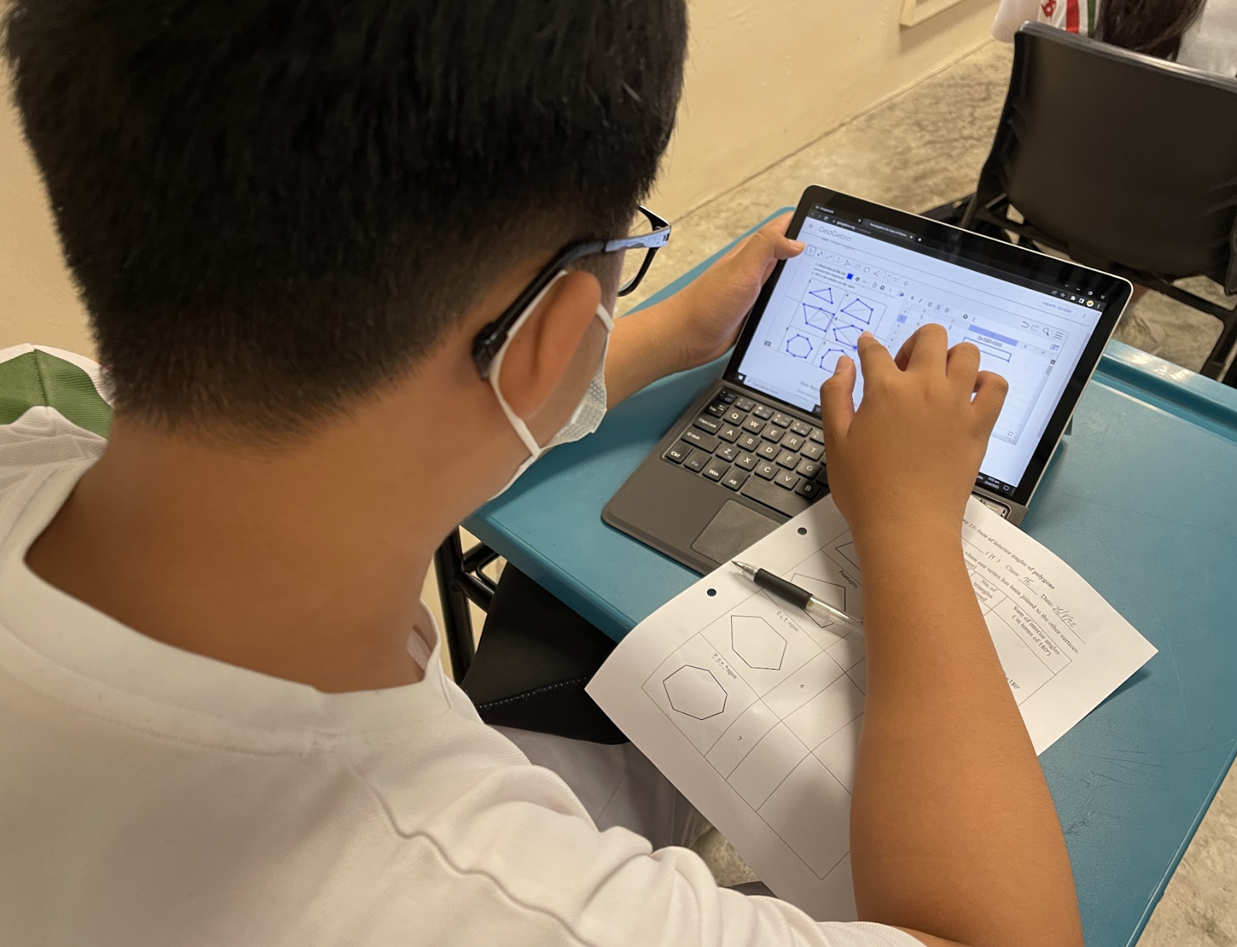 A masked boy with glasses touches a tablet showing polygon shapes, with a geometry worksheet on the desk.