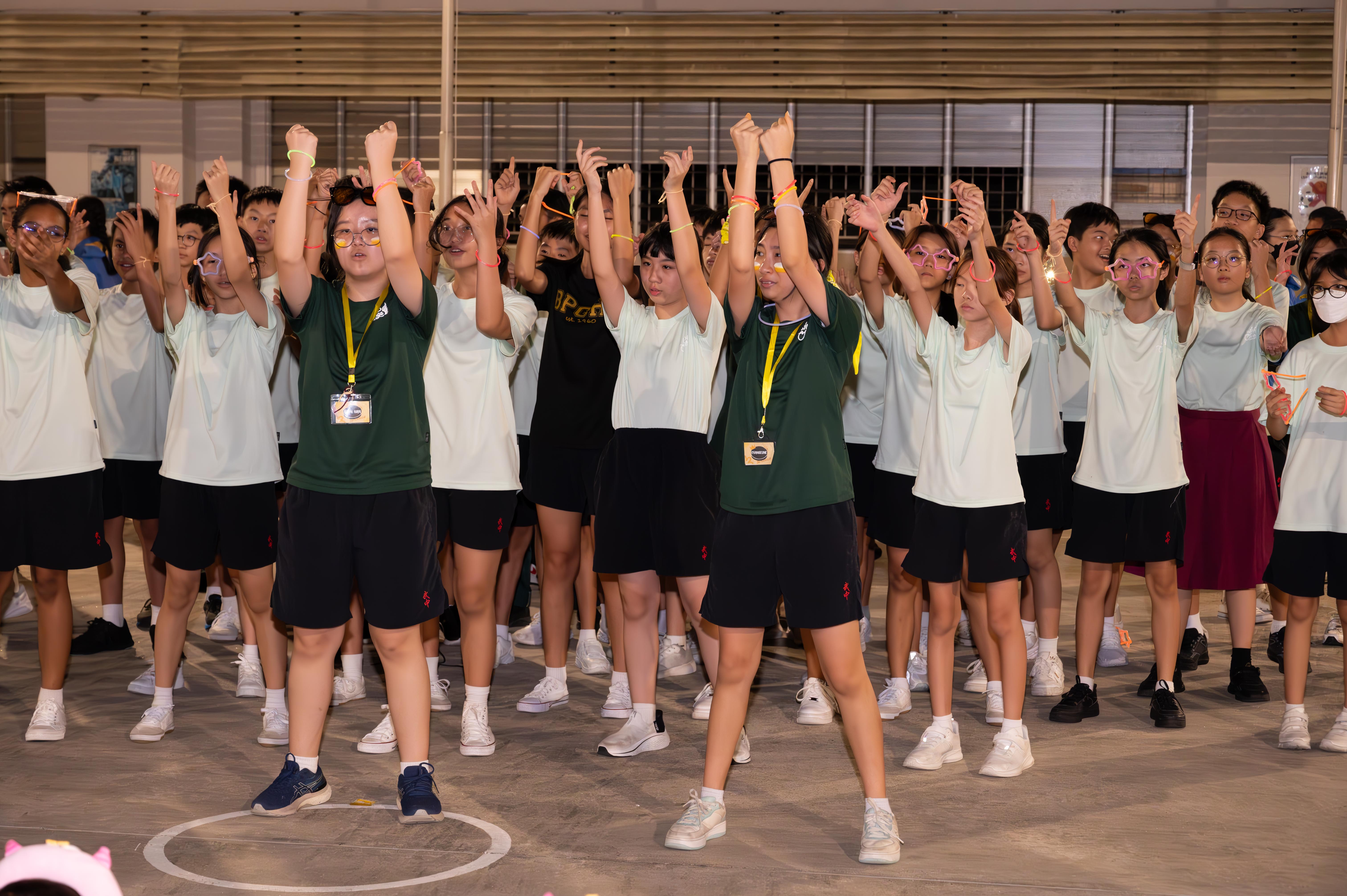 A large group of students in matching uniforms raise their arms during an indoor school event.