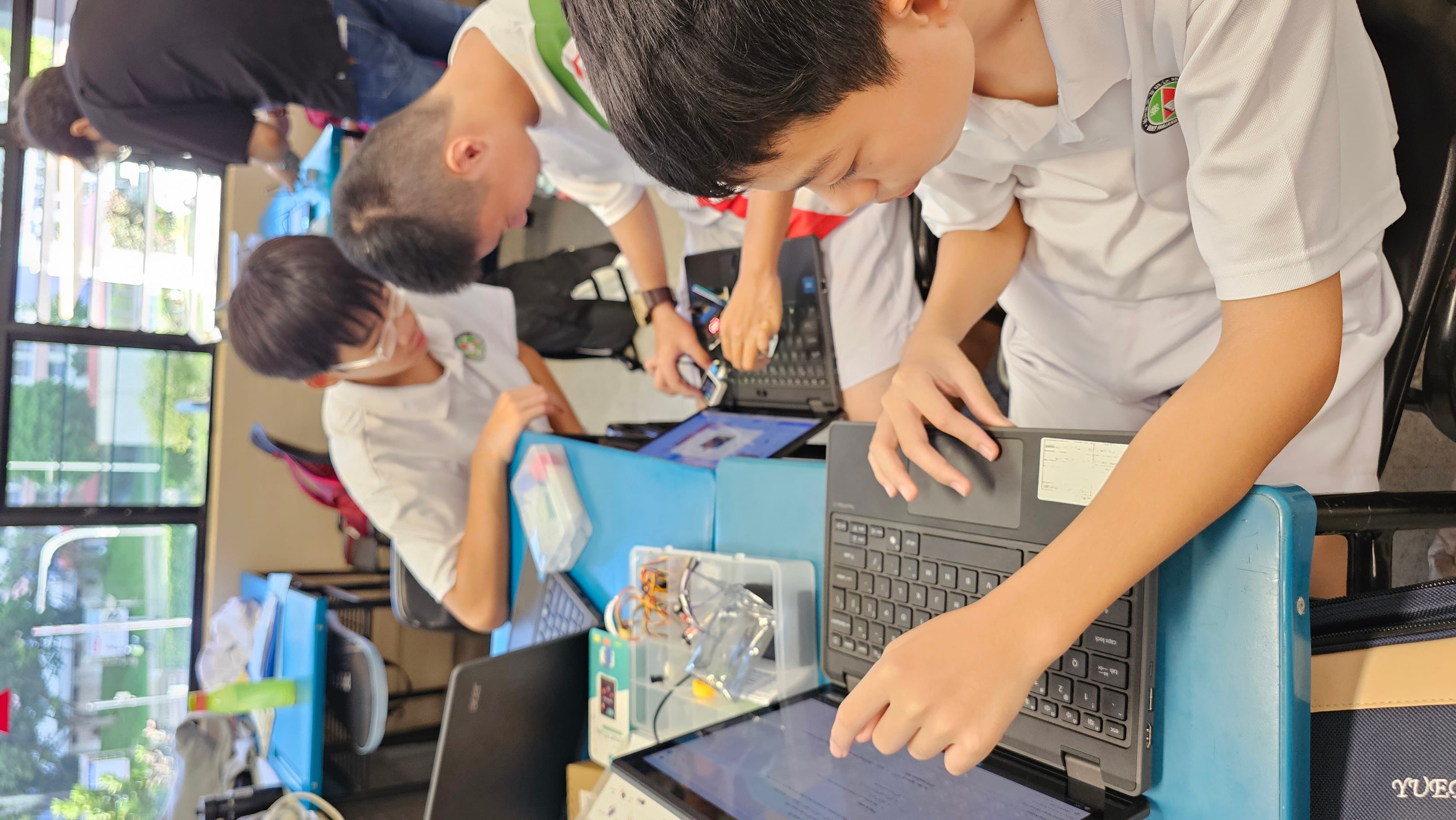Three students in white uniforms work on laptops at blue desks in a classroom with electronics components nearby.