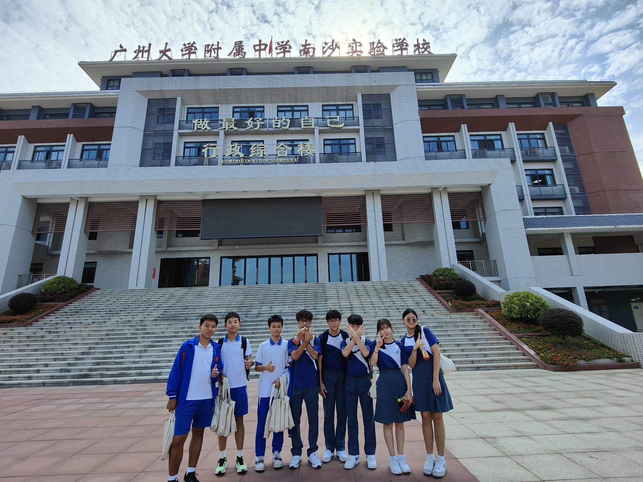 Seven students in blue and white uniforms pose in front of a large Chinese school administration building with steps.