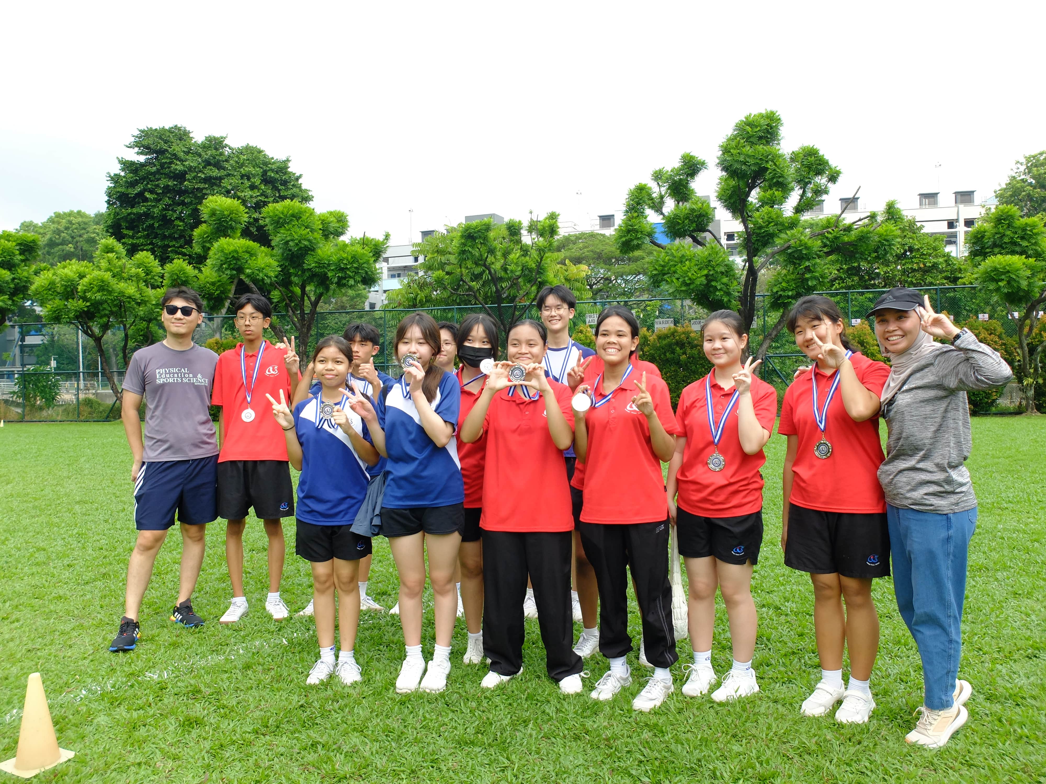 A group of students and two adults pose on a grass field, wearing medals and making peace signs.