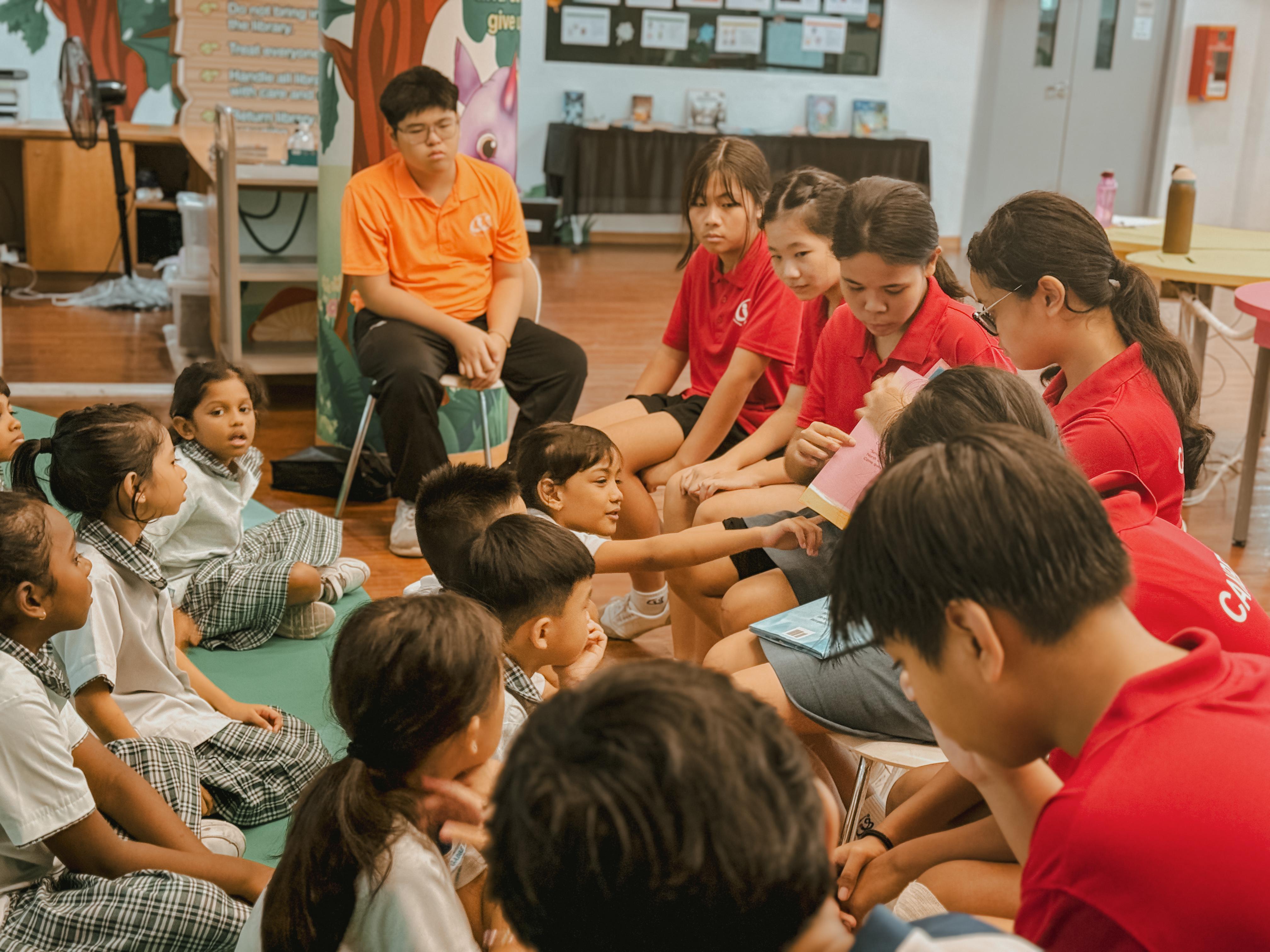 A group of secondary school students wearing red tees read a book to a group of primary school students sitting on the floor in front of them, as a young girl points to something in the book