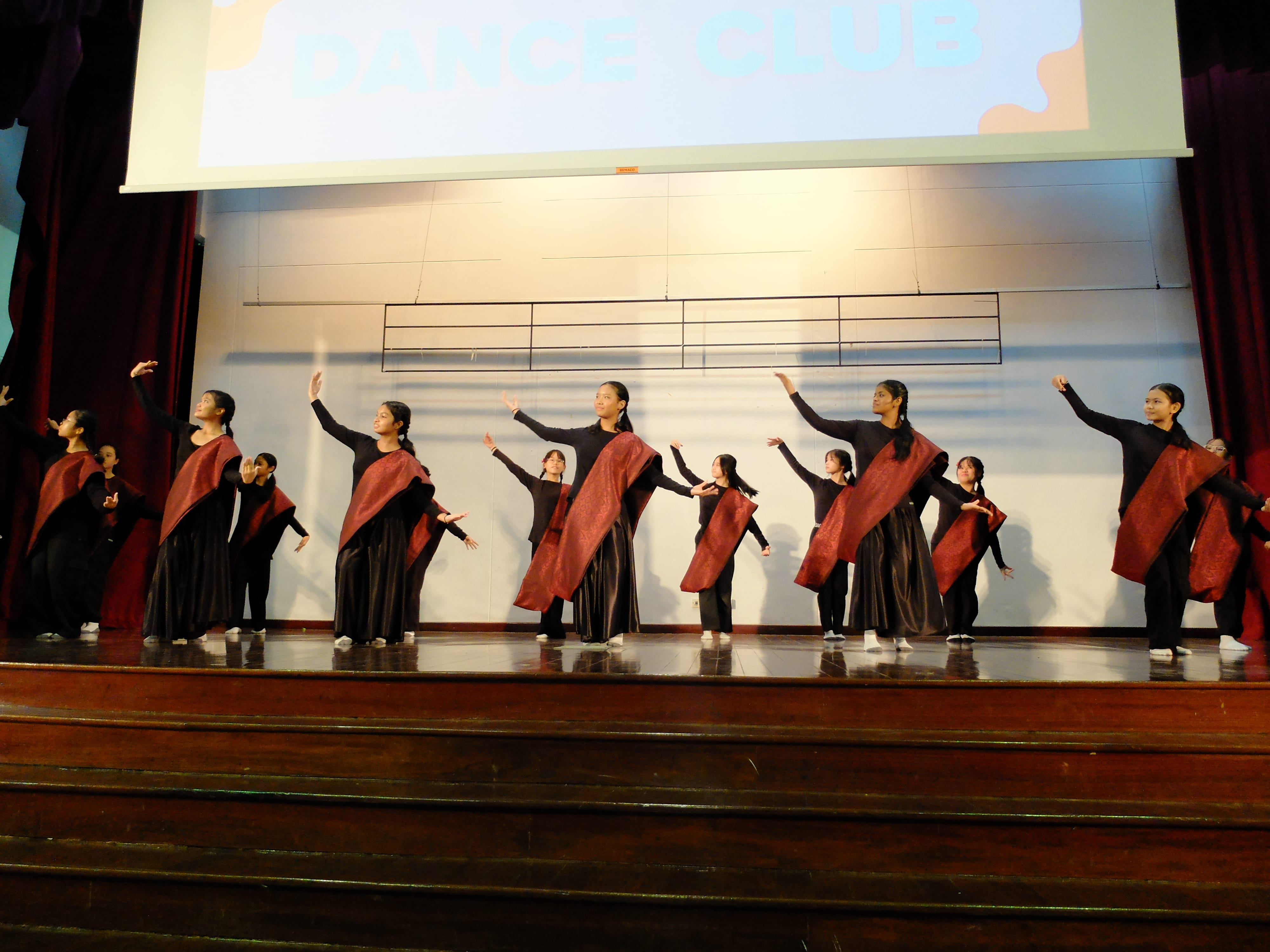 Girls in black and red costumes perform a synchronized dance on a stage under a Dance Club banner.